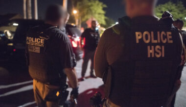 Two Department of Homeland Security agents face away from the camera outdoors at night