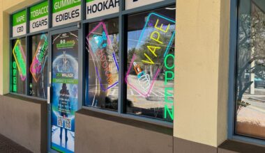 Neon signs and other signs in the store window of a store appear next to the sidewalk at a vape shop near Windermere.