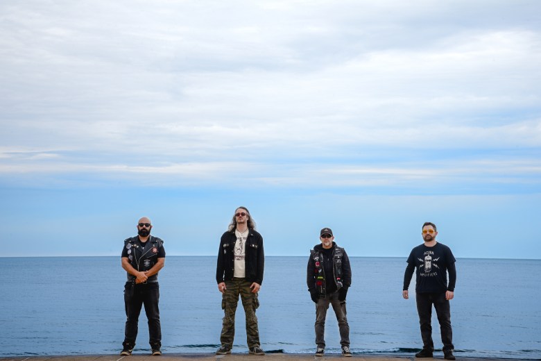 Four people stand evenly spaced in a horizontal line on a stone ledge overlooking a vast, calm body of water under an expansive, cloudy blue sky. They are all wearing dark, casual clothing, including t-shirts and jackets, with three of the four wearing sunglasses. The individual on the far left has a full beard and a leather vest with patches, while the individual second from the left has long wavy hair and camouflage pants.