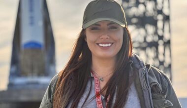 Woman wearing olive green baseball cap smiles as she stands in front of New Glenn rocket