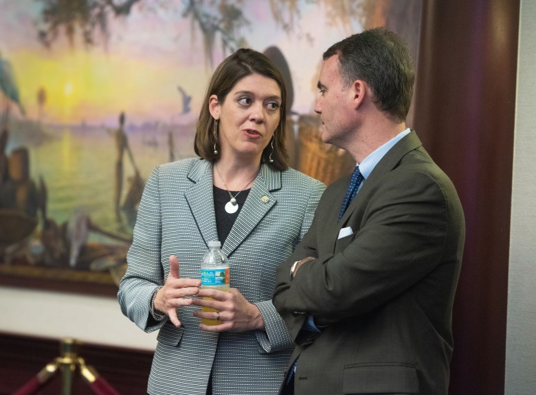 Two people in professional attire stand in conversation indoors. The individual on the left is wearing a grey textured blazer, a silver pendant necklace, and long earrings while holding a plastic water bottle and gesturing with their hand. The individual on the right is wearing a dark olive suit jacket and a blue patterned tie, standing with their arms crossed. In the background, a large landscape painting of a sunset over a body of water is partially visible.