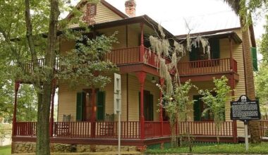 The John G. Riley House, a historic two-story wood-frame home with wraparound porches, red railings, green shutters, and surrounding trees.