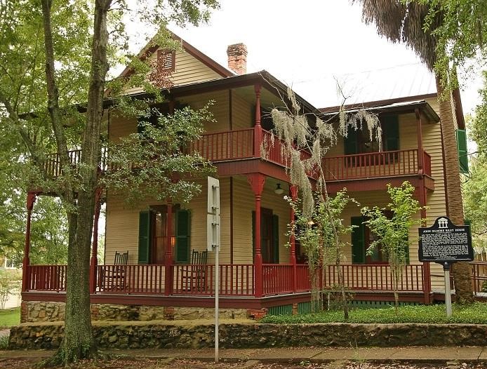 The John G. Riley House, a historic two-story wood-frame home with wraparound porches, red railings, green shutters, and surrounding trees.