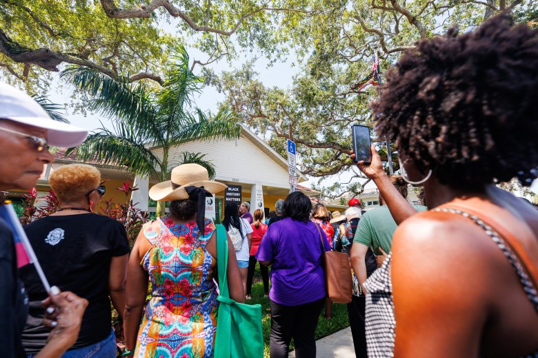 A view from behind a crowd of people gathered outdoors for an event at a white, single-story building. In the center, a person stands on a porch next to a black sign that reads "BLACK HISTORY MATTERS." Attendees in the foreground wear colorful summer clothing, including a bright patterned dress and a purple t-shirt, while one person on the right holds up a smartphone to record the scene. The setting is lush with large oak trees and palm trees under a clear, sunny sky.