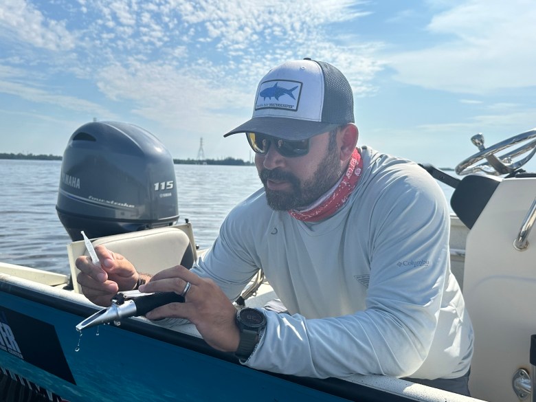 A close-up shot of a person with a beard wearing a grey long-sleeved shirt, a red patterned neck gaiter, sunglasses, and a "Tampa Bay Waterkeeper" baseball cap. They are seated on a boat and focused on using a handheld scientific instrument, likely a refractometer, while holding a small plastic dropper. The background shows a Yamaha outboard motor and a calm body of water under a bright, wispy sky.
