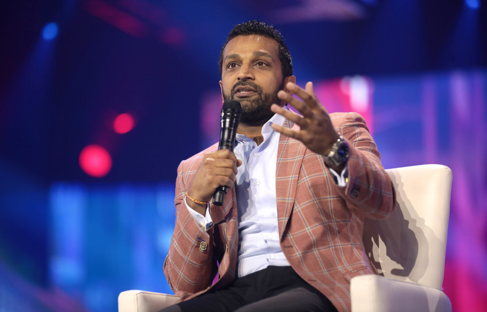 Kash Patel wears a pink and blue plaid sports jacket sitting in a white chair. He is speaking holding a microphone in his right hand and he is gesturing with his left hand extended. He is wearing a light blue button down and black pants.