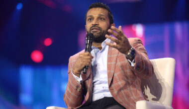 Kash Patel wears a pink and blue plaid sports jacket sitting in a white chair. He is speaking holding a microphone in his right hand and he is gesturing with his left hand extended. He is wearing a light blue button down and black pants.