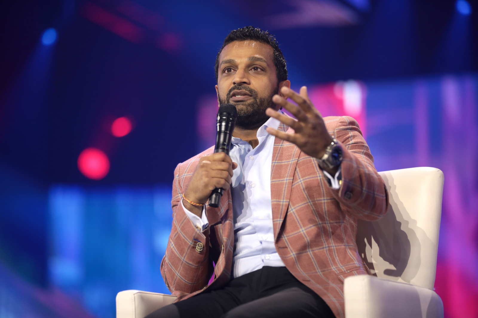 Kash Patel wears a pink and blue plaid sports jacket sitting in a white chair. He is speaking holding a microphone in his right hand and he is gesturing with his left hand extended. He is wearing a light blue button down and black pants.