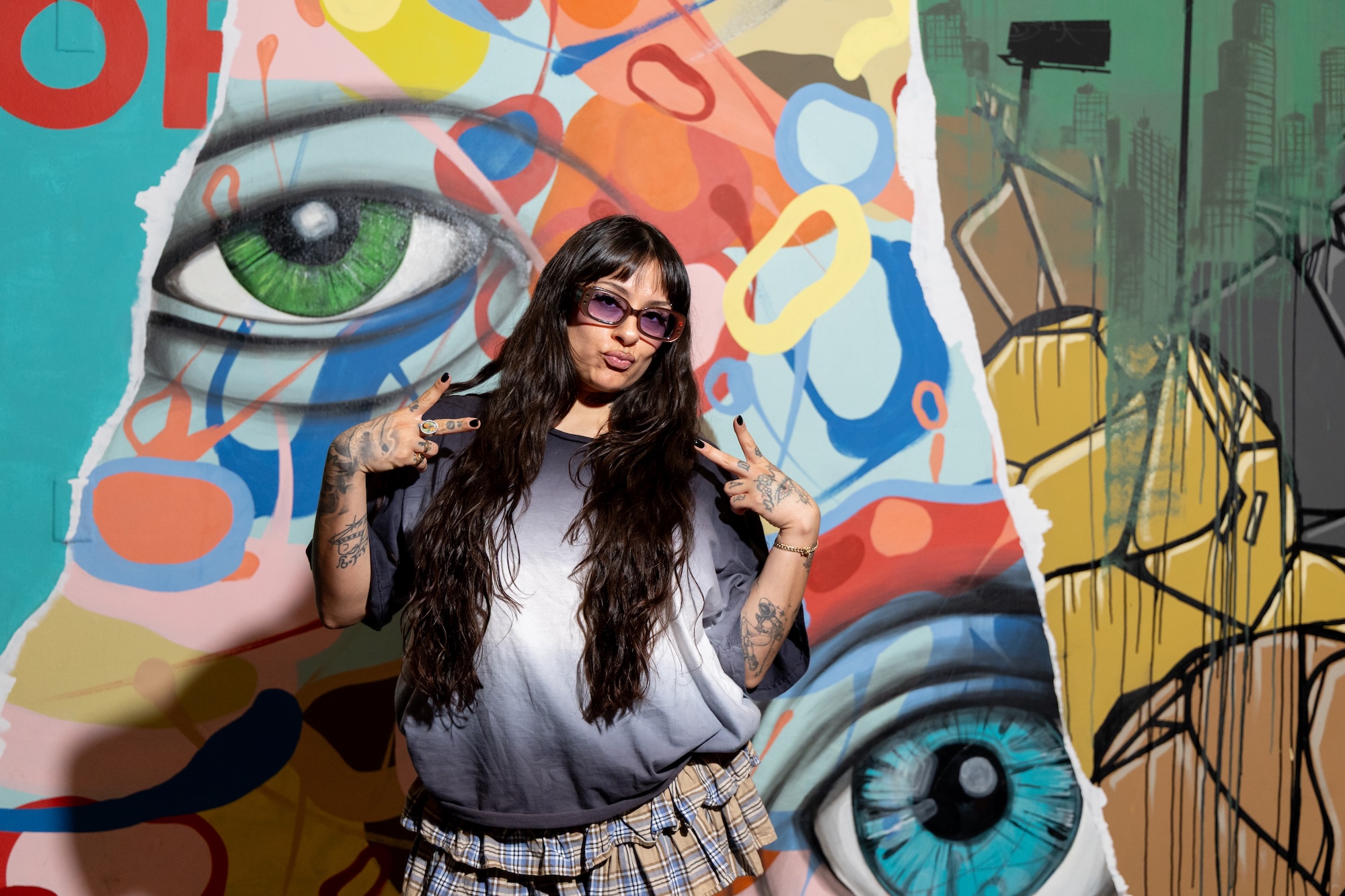 photo of a woman in a baggy shirt posing in front of a mural with painted eyes and splotches of paint
