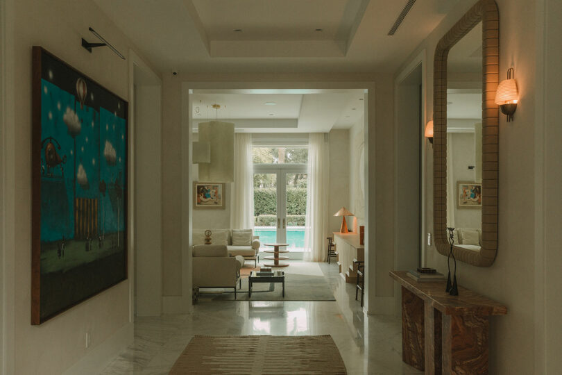 A hallway with a large mirror and painting leads to a living room with beige furniture and glass doors opening to a backyard with a swimming pool.