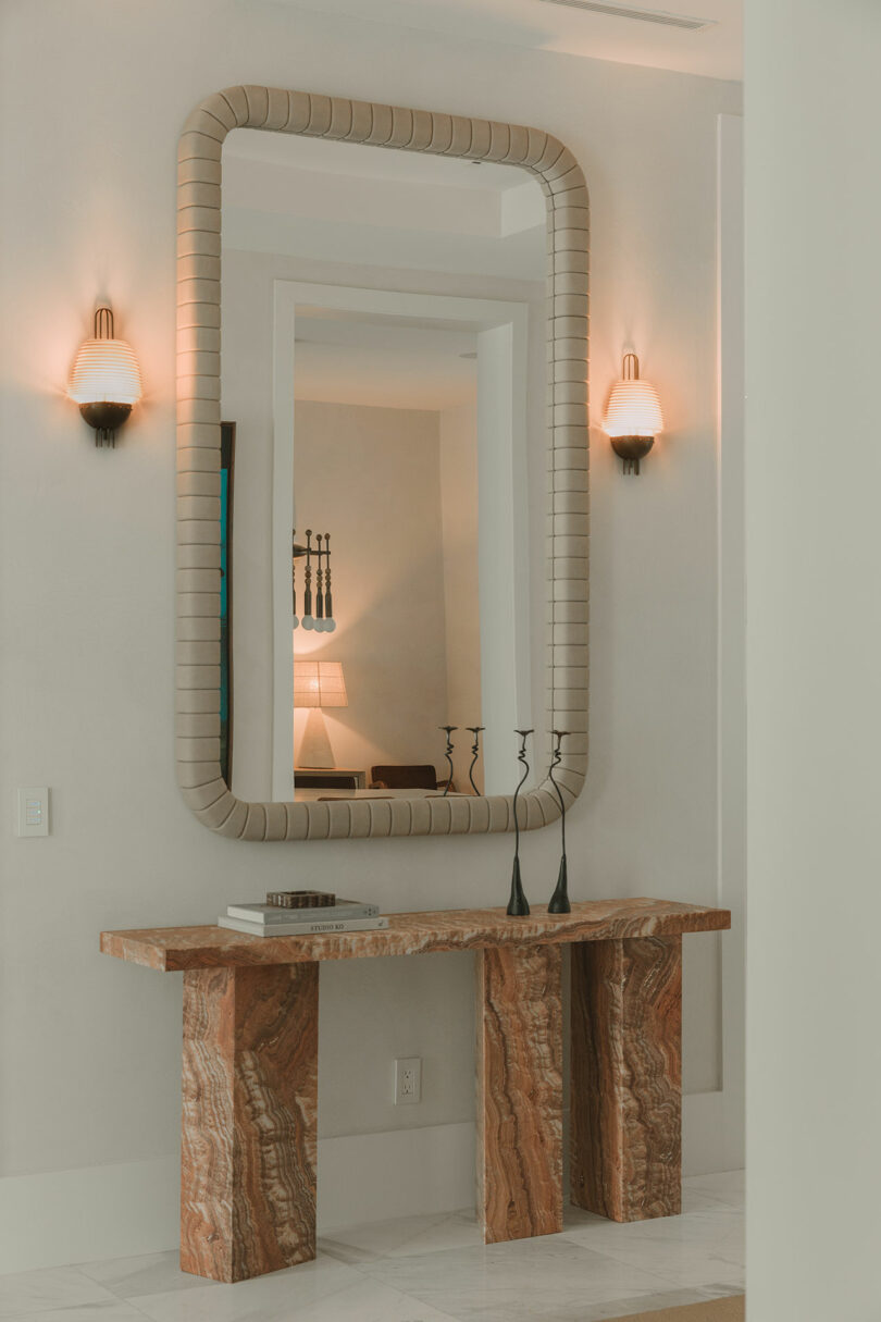 A stone console table with books and candle holders sits below a large rectangular mirror, flanked by two wall sconces in a neutral-toned interior hallway.