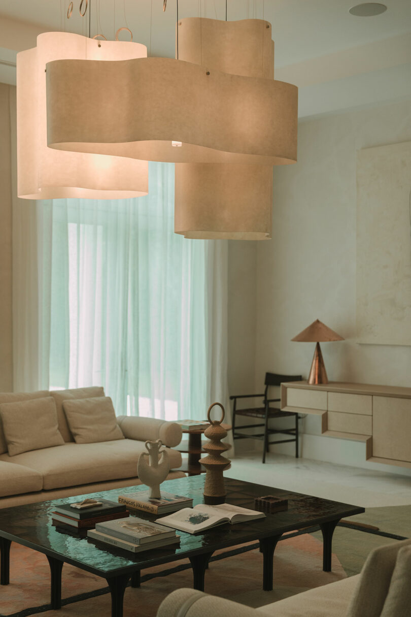 Modern living room with beige sofas, a black coffee table with books and decor, large sculptural pendant lights, sheer curtains, and minimalist furniture.