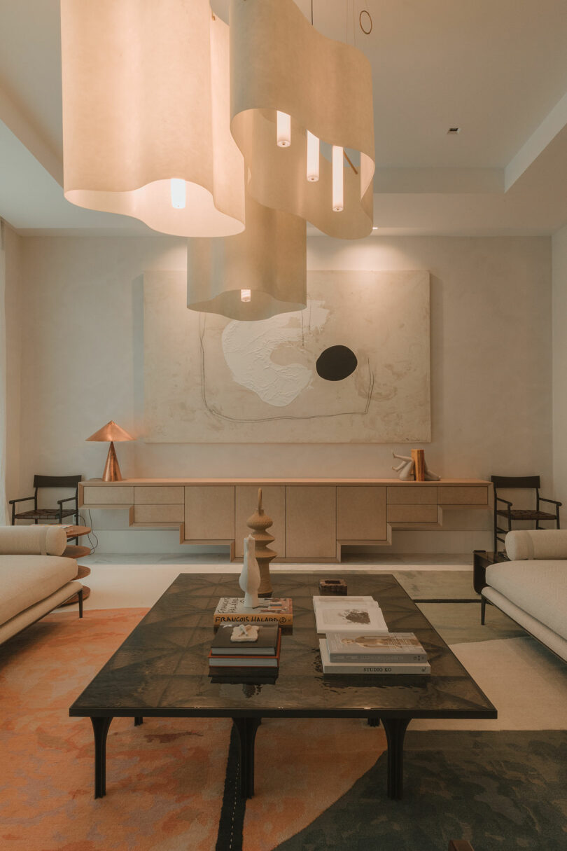 A modern living room with two sofas, a black coffee table with decor and books, pendant lights, abstract wall art, and a sideboard with decorative objects.