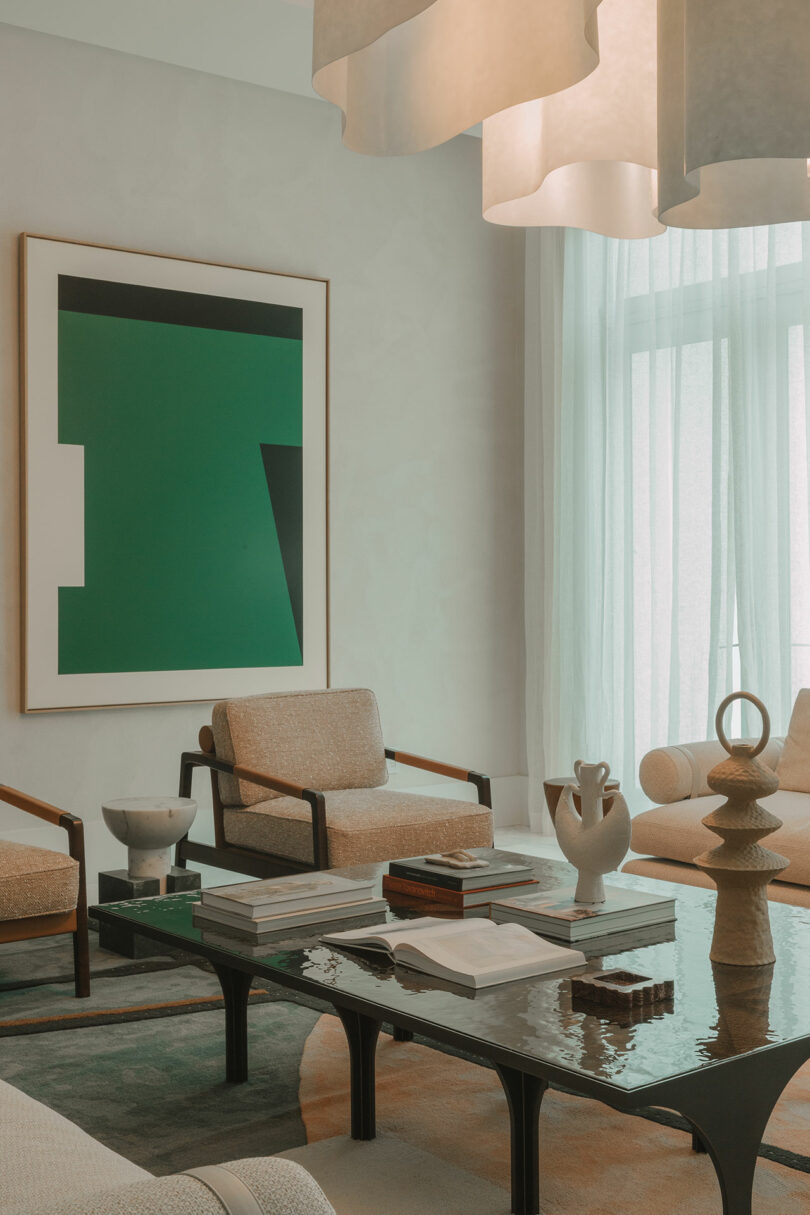 A modern living room with neutral-toned furniture, a glass coffee table holding books and ceramics, large abstract artwork on the wall, and sheer curtains covering a window.
