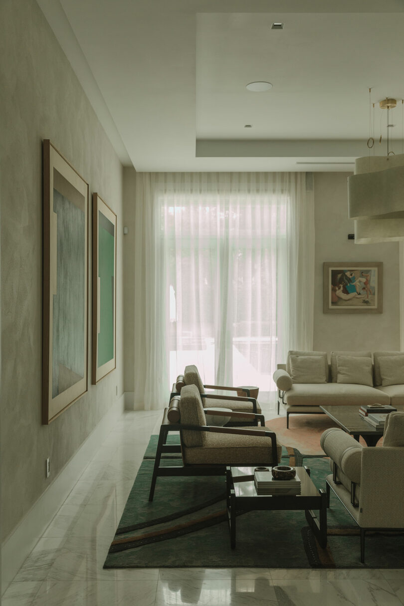Modern living room with beige armchairs, a cream sofa, coffee tables, abstract wall art, and sheer curtains letting in natural light.
