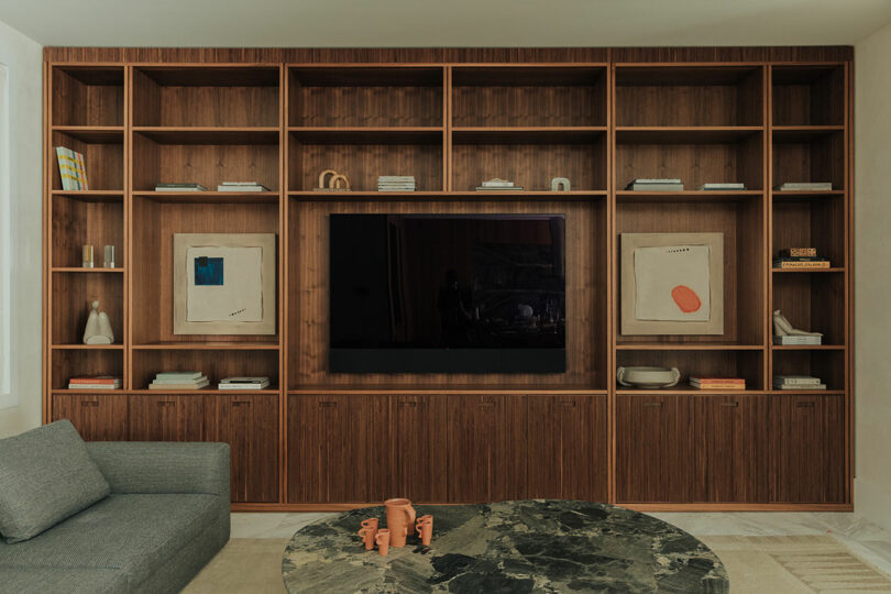 A wooden built-in shelving unit with a TV at the center, books, art, and decorative objects displayed on shelves, beside a gray sofa and a round marble coffee table.