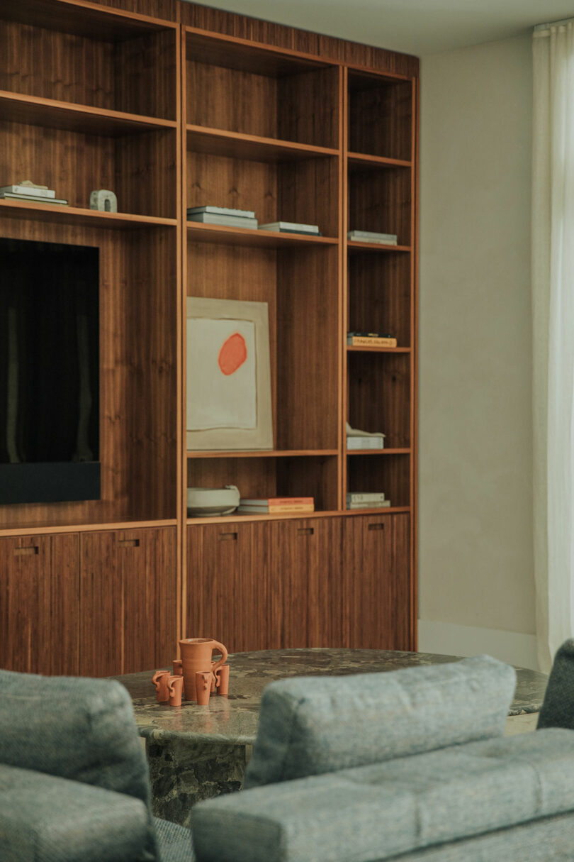 A wooden built-in bookshelf with sparse decor items, books, and art, next to a stone coffee table with terra cotta cups, and gray upholstered chairs in the foreground.