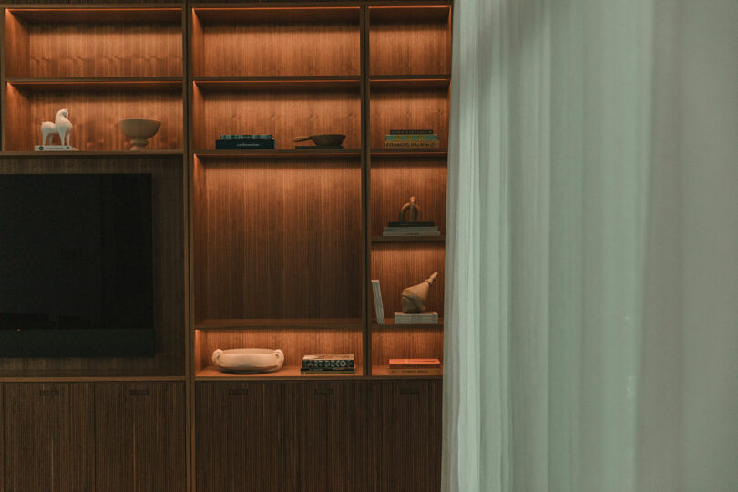 Wooden shelves with built-in lighting display books, decor items, and sculptures; a portion of a TV and sheer curtains are also visible.