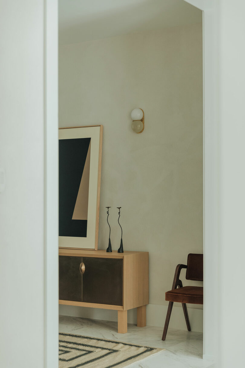 Minimalist interior with a wooden cabinet, abstract framed artwork, two black candlesticks, a wall sconce, and a wooden chair with a dark seat.