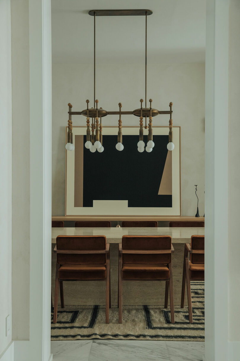 A modern dining room with a long wooden table, eight matching wooden chairs, a geometric black and beige artwork, and a chandelier with exposed bulbs.