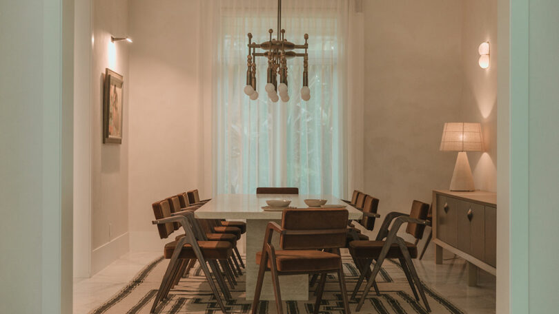 A modern dining room with a rectangular table, eight brown chairs, a chandelier, sideboard with a lamp, and sheer curtains covering tall windows.