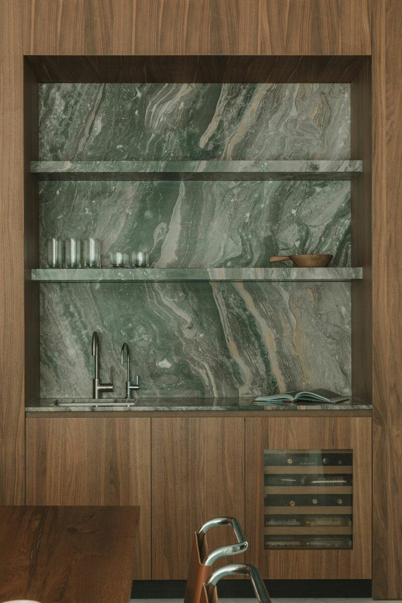 A modern kitchen nook with green marble backsplash, wood cabinets, glass shelves holding glasses and bowls, a sink, an open book, and a small wine fridge.