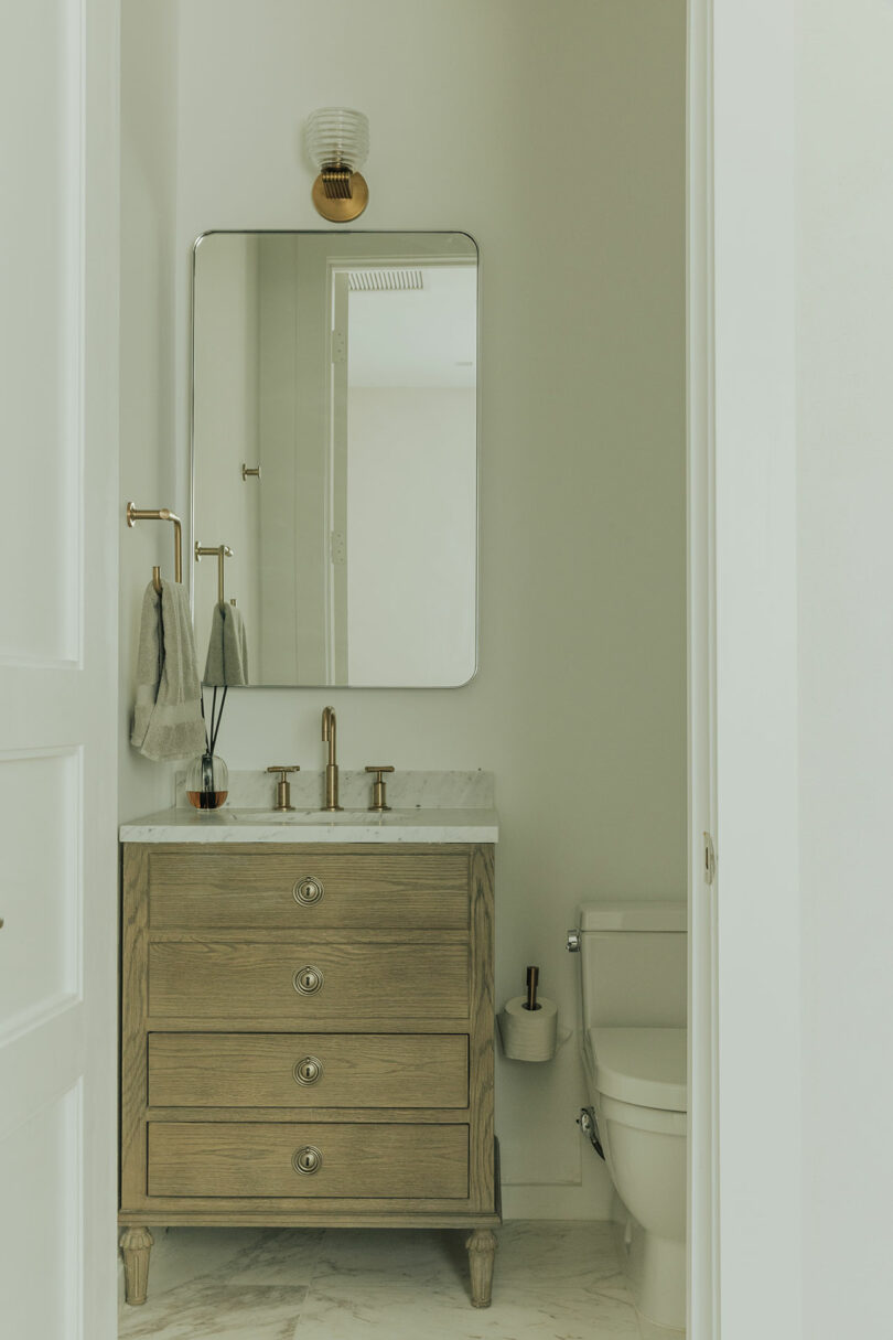Small bathroom with a wooden vanity, marble countertop, rectangular mirror, wall sconce, towel ring, and toilet on a light marble floor.