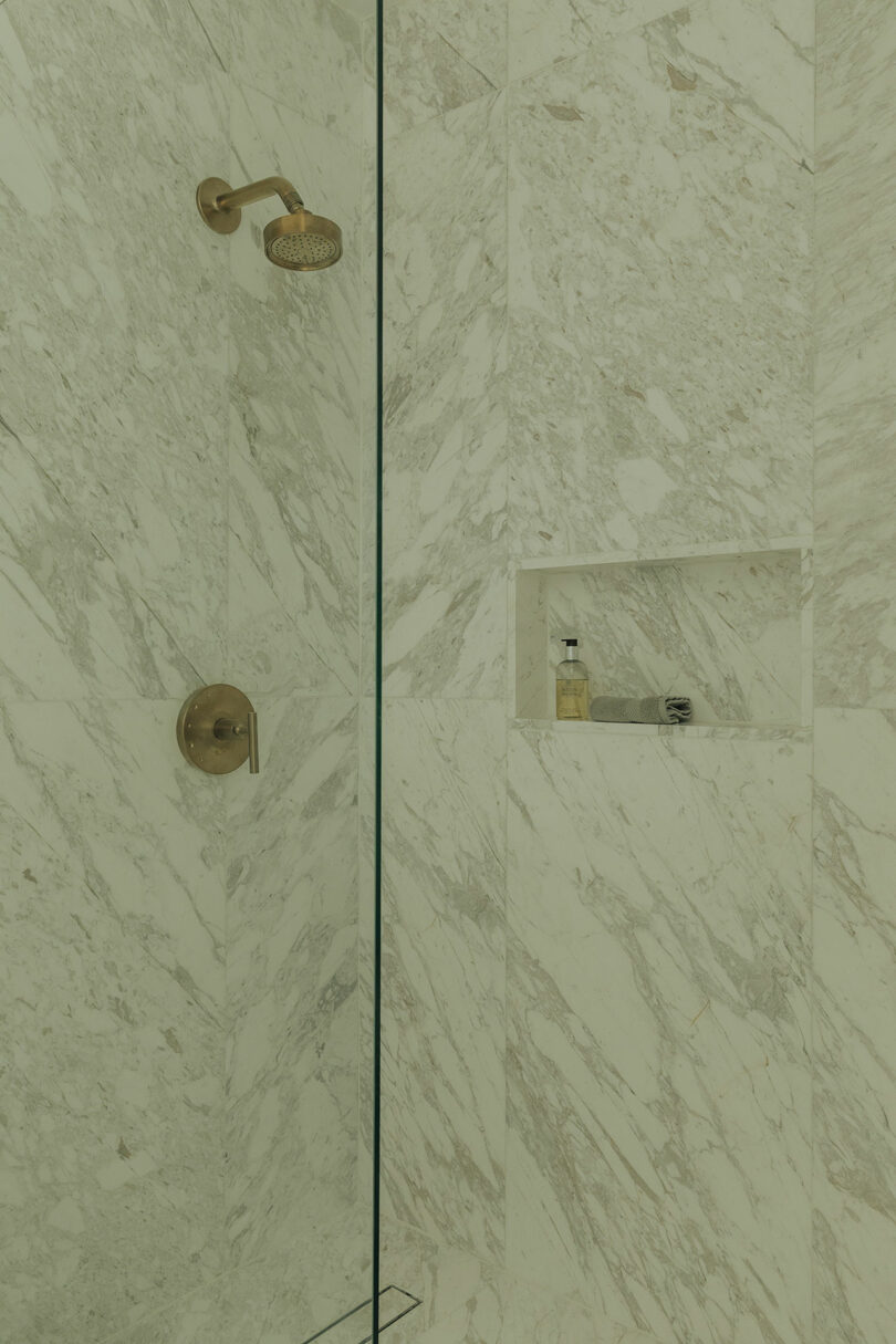 A marble-tiled shower with a brass showerhead and control, and a built-in niche holding a bottle and a bar of soap.