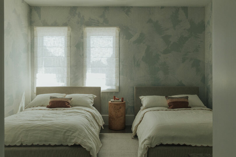 A bedroom with two double beds, a shared wooden nightstand between them, and two windows with sheer curtains letting in natural light.