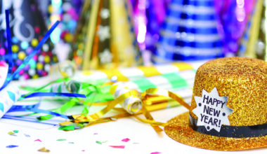 Gold glitter top hat with a “Happy New Year!” label sits on a table surrounded by colorful party hats, streamers, and confetti, suggesting a New Year’s celebration.