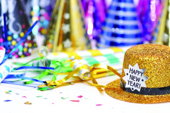 Gold glitter top hat with a “Happy New Year!” label sits on a table surrounded by colorful party hats, streamers, and confetti, suggesting a New Year’s celebration.