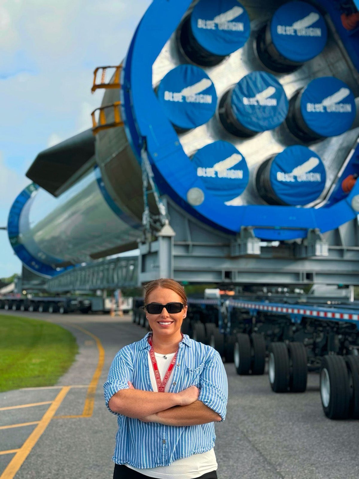 Woman in light blue shirt stands with arms crossed in front of Blue Origin rocket on transporter vehicle