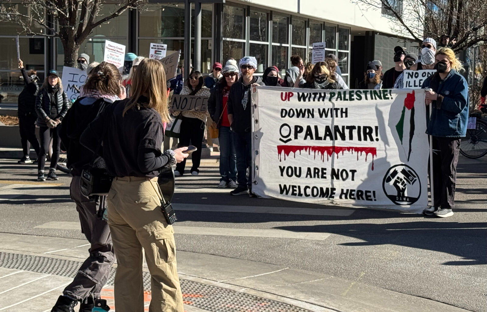 Protesters hold up a canvas sign in front of two people and that says says "Up with Palestine down with Palantir! You are not welcome here. There is a line of blood splatter under Palantir. There is also a drawing of the outline of Israel with a palestine flag drawn vertically over the outline. There is a fist at the bottom right corner.