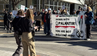Protesters hold up a canvas sign in front of two people and that says says "Up with Palestine down with Palantir! You are not welcome here. There is a line of blood splatter under Palantir. There is also a drawing of the outline of Israel with a palestine flag drawn vertically over the outline. There is a fist at the bottom right corner.