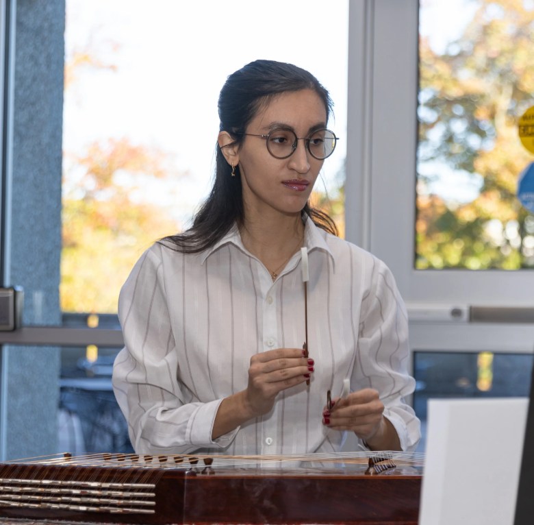 Musician Parastoo Shafiei performs on the santur, a hammered dulcimer. She is wearing a white pinstripe shirt and round glasses, holding two delicate wooden mallets (mezrabs) over the strings of the traditional instrument in a brightly lit indoor setting.
