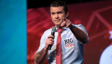 Pete Hegseth holding a microphone in his right hand and pointing with his left. He is wearing a blue button down shirt with the sleeves rolled up. He has a red tie and has a stick on the left side of his chest that says "BIG government Sucks." He has a tattoo on his right arm that says "We the People."