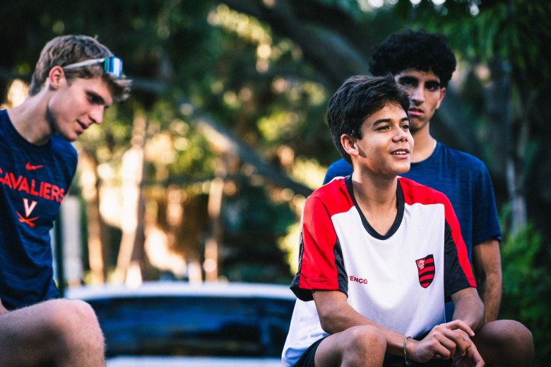 Plant High School runner Igor Gordiano in a red and white jersey sitting with teammates at Fred Ball Park in Tampa after a practice session with Meb Keflezighi.