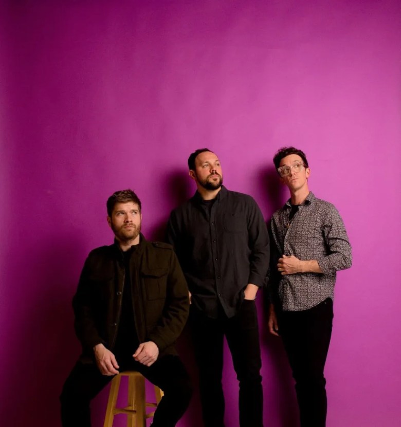 Three people stand against a vibrant, solid purple background. The individual on the left is seated on a wooden stool, wearing a dark olive jacket and looking off-camera. The individual in the center stands with hands in their pockets, wearing a black button-down shirt and looking upward. The individual on the right stands in a patterned grey-and-white shirt and clear-framed glasses, also looking upward with one hand near their chest.