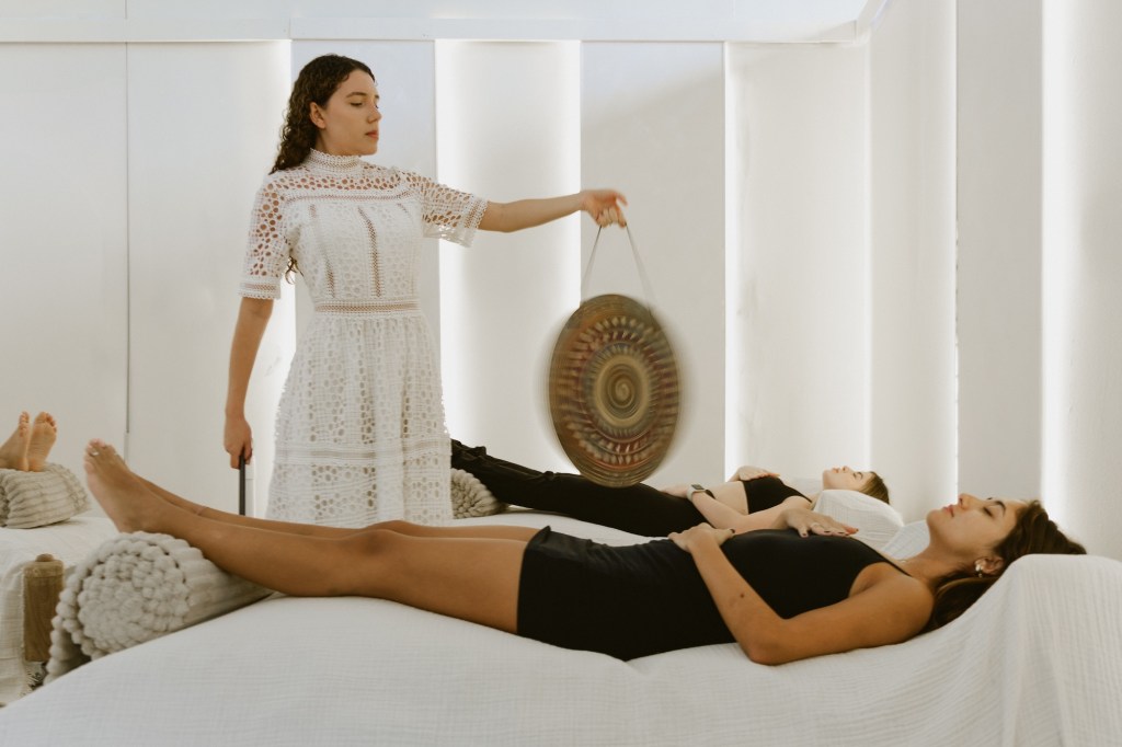 photo of a woman in a white dress playing a gong inside a wellness studio as two women lie down on beds on either sides of her