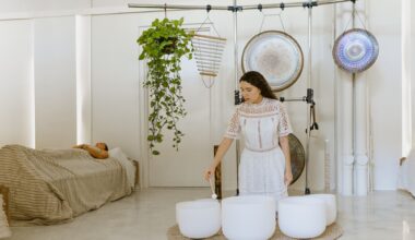 Photo of two women inside a wellness studio. The woman on the left lies in bed covered in blankets. The woman on the right plays three sound bowls as gongs, chimes, and a plant hang behind her