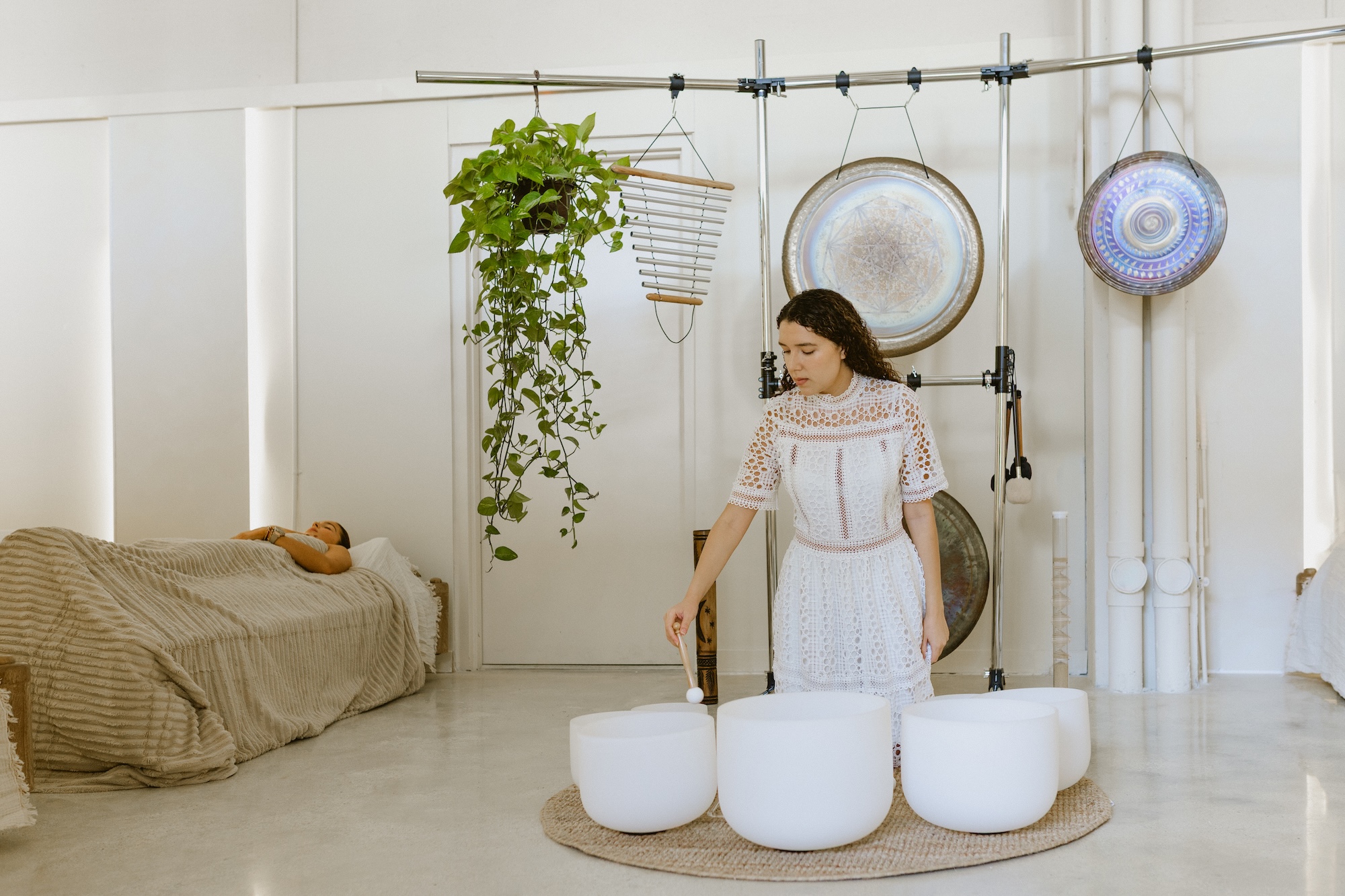 Photo of two women inside a wellness studio. The woman on the left lies in bed covered in blankets. The woman on the right plays three sound bowls as gongs, chimes, and a plant hang behind her