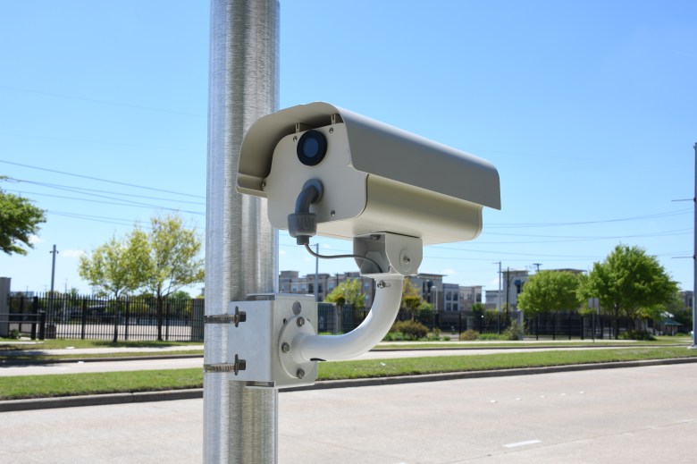 Close-up of a RedSpeed speed and red-light enforcement camera mounted on a metal pole against a clear blue sky, showing the high-resolution lens and protective housing used for automated traffic enforcement.