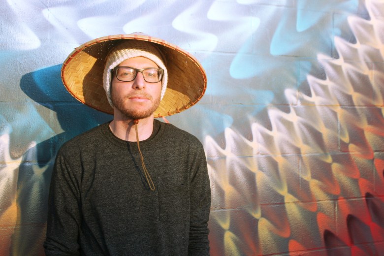 Tampa Bay producer and percussionist Roger Thomas posing for a portrait by Mike Nyman. He wears a conical straw hat over a white knit beanie and glasses, standing against a colorful graffiti wall with wavy blue, yellow, and red patterns.