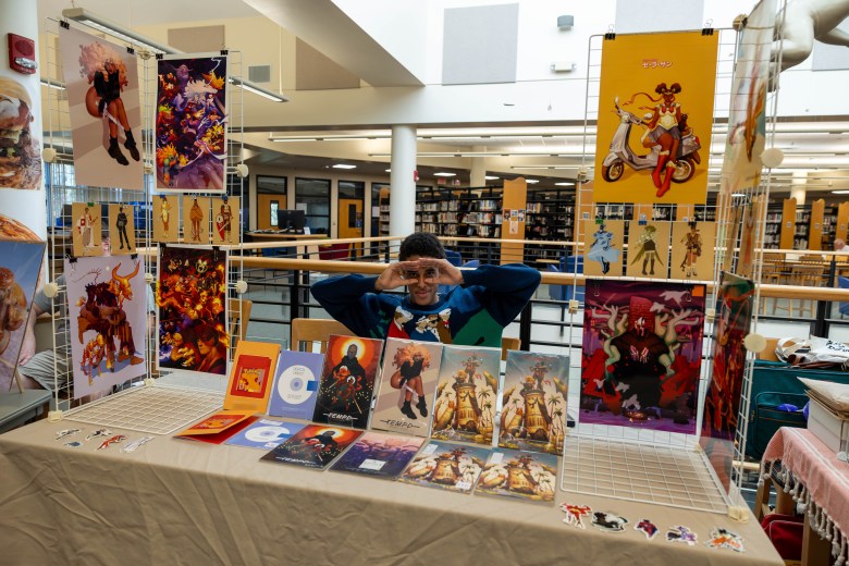A young artist posing playfully behind a table filled with original prints and zines at a library for St. Pete Zine Fest.