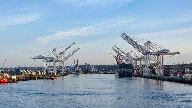 A wide landscape view of a busy shipping terminal waterway under a pale blue, late-afternoon sky. Several massive white container cranes line both sides of the deep blue water, with a large cargo ship filled with colorful shipping containers docked on the right. In the distance, the shoreline features industrial buildings and rolling hills, while the foreground shows the gentle wake of a vessel on the water's surface.