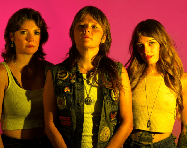 A promotional portrait of three women in the band Thelma and the Sleaze posing confidently against a bright pink background