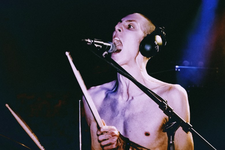 A high-energy, close-up shot of a shirtless drummer performing on a dark stage. The individual has a shaved head, is wearing large black headphones, and is shouting into a microphone with a wide-mouthed expression of intense focus. They are holding wooden drumsticks mid-performance, with the scene illuminated by a dramatic blue spotlight in the upper right corner.