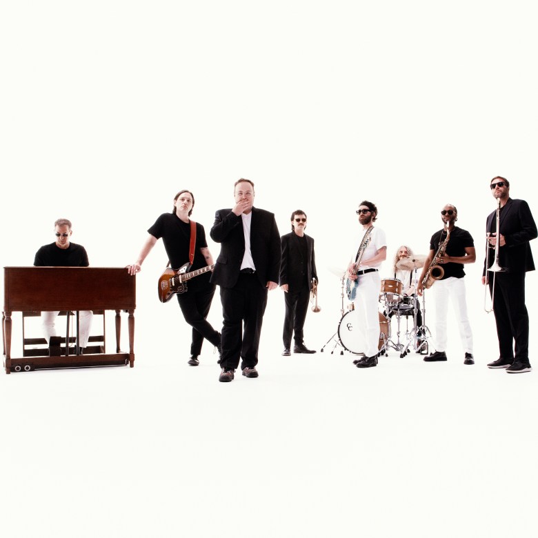 St. Paul and The Broken Bones band members posing in a minimalist, high-key studio setting. The eight-piece soul band is dressed in a mix of black and white outfits with musical instruments including a soul-style organ, electric guitar, saxophone, and trumpet against a solid white background.