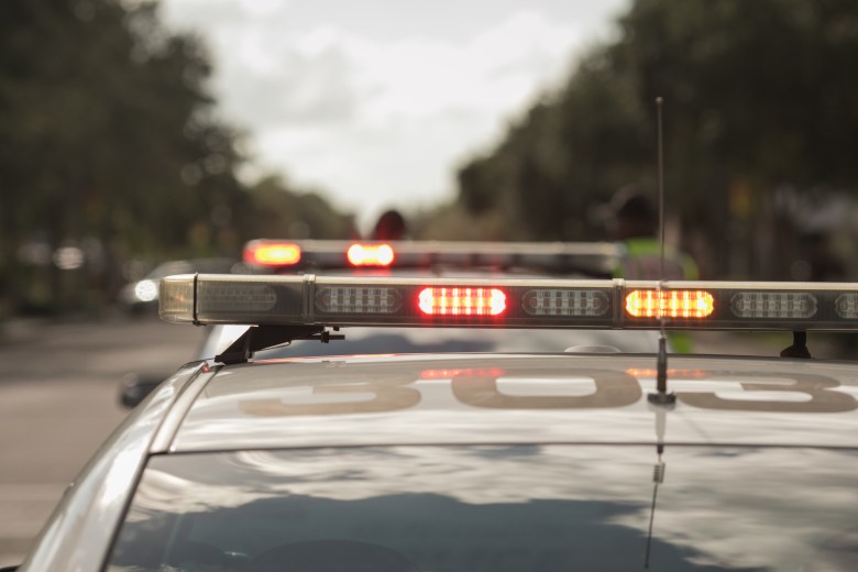 A close-up, low-angle shot from behind a police cruiser, focusing on the illuminated red and amber emergency light bar on the roof. The numbers "303" are visible on the trunk in the foreground. In the blurred background, another police vehicle's lights are visible on a tree-lined street under a bright, overcast sky.