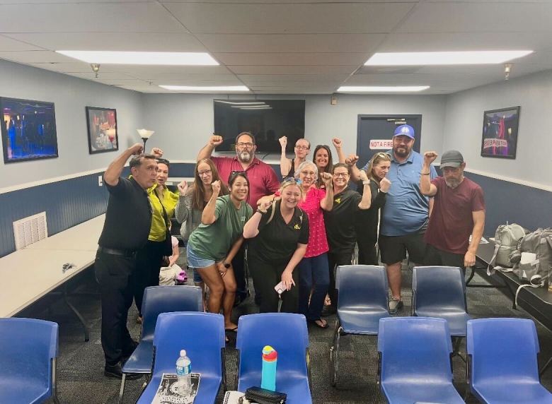 A group of thirteen people stand together in a breakroom or meeting space, all raising one fist in a gesture of solidarity. They are dressed in casual and work attire, including t-shirts and polo shirts, smiling toward the camera. The room has grey walls with blue wainscoting, several rows of blue plastic chairs in the foreground, and long white tables along the side.